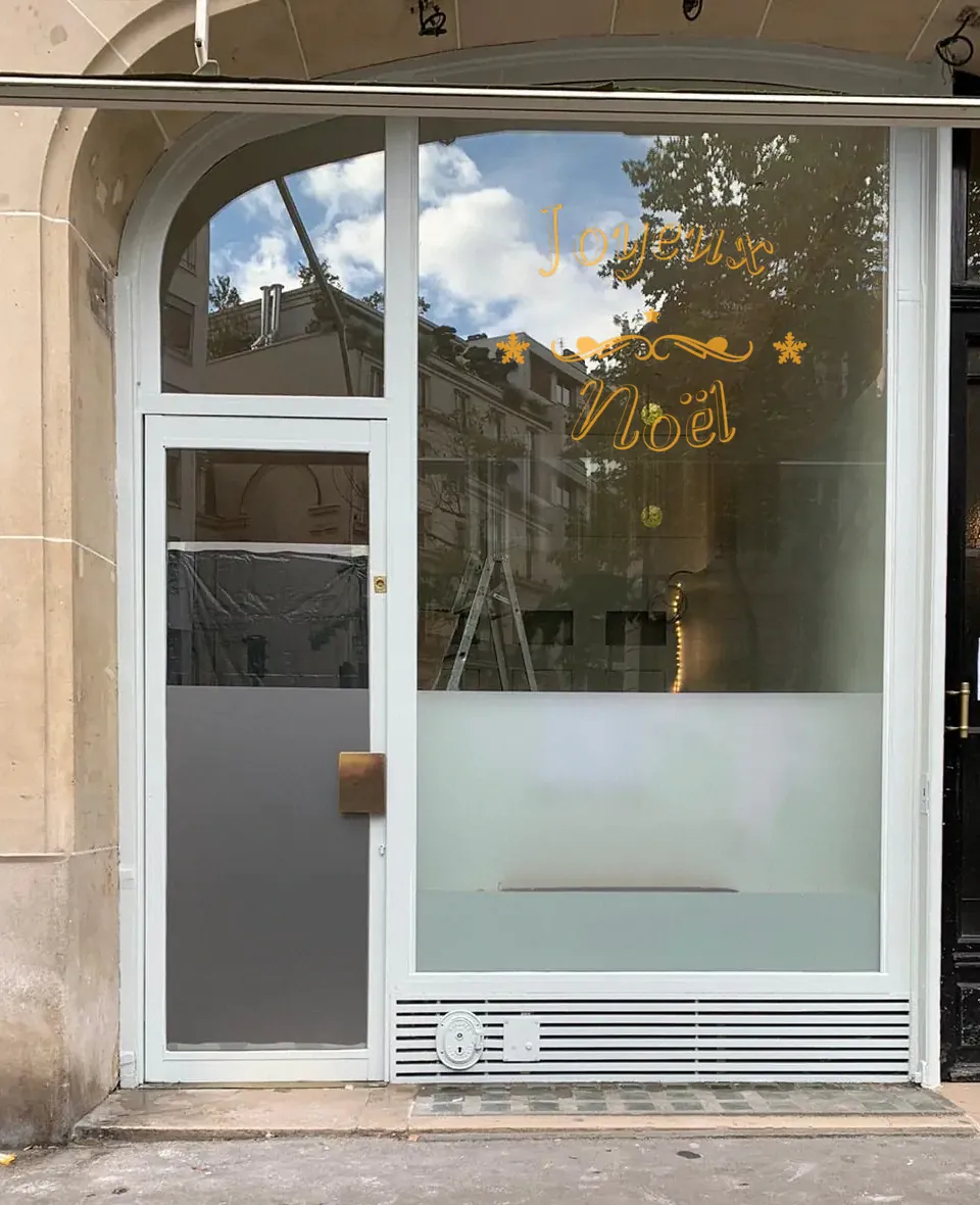 Lettrage « Joyeux Noël » calligraphié au trait doré avec arabesque et flocons, adhésif découpé sur vitrine.