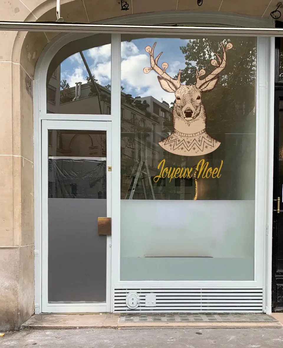 Tête de cerf au pull de Noël en vitrophanie imprimée et découpée, posée sur une vitrine avec le lettrage « Joyeux Noël » manuscrit doré.