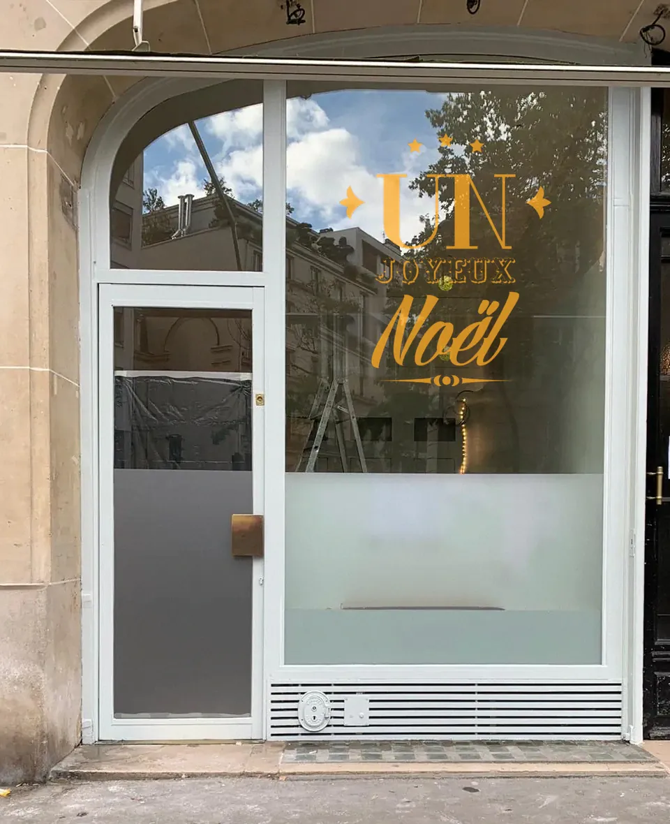 Composition typographique « Un Joyeux Noël » dorée en adhésif découpé, posée sur une grande vitrine.