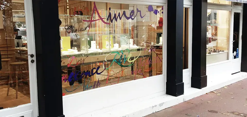 Boutique Arthus-Bertrand pour les fêtes : lettrage manuscrit multicolore « Aimer » en vitrophanie sur toute la longueur de la vitrine.