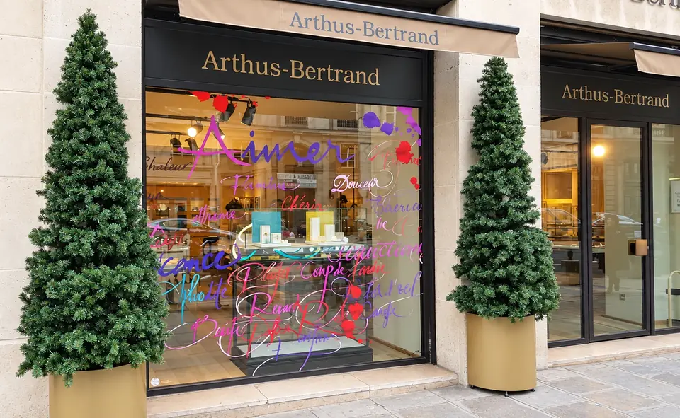 Façade de la boutique Arthus-Bertrand pour les fêtes : lettrage manuscrit coloré en vitrophanie sur la vitrine, encadrée de deux sapins.