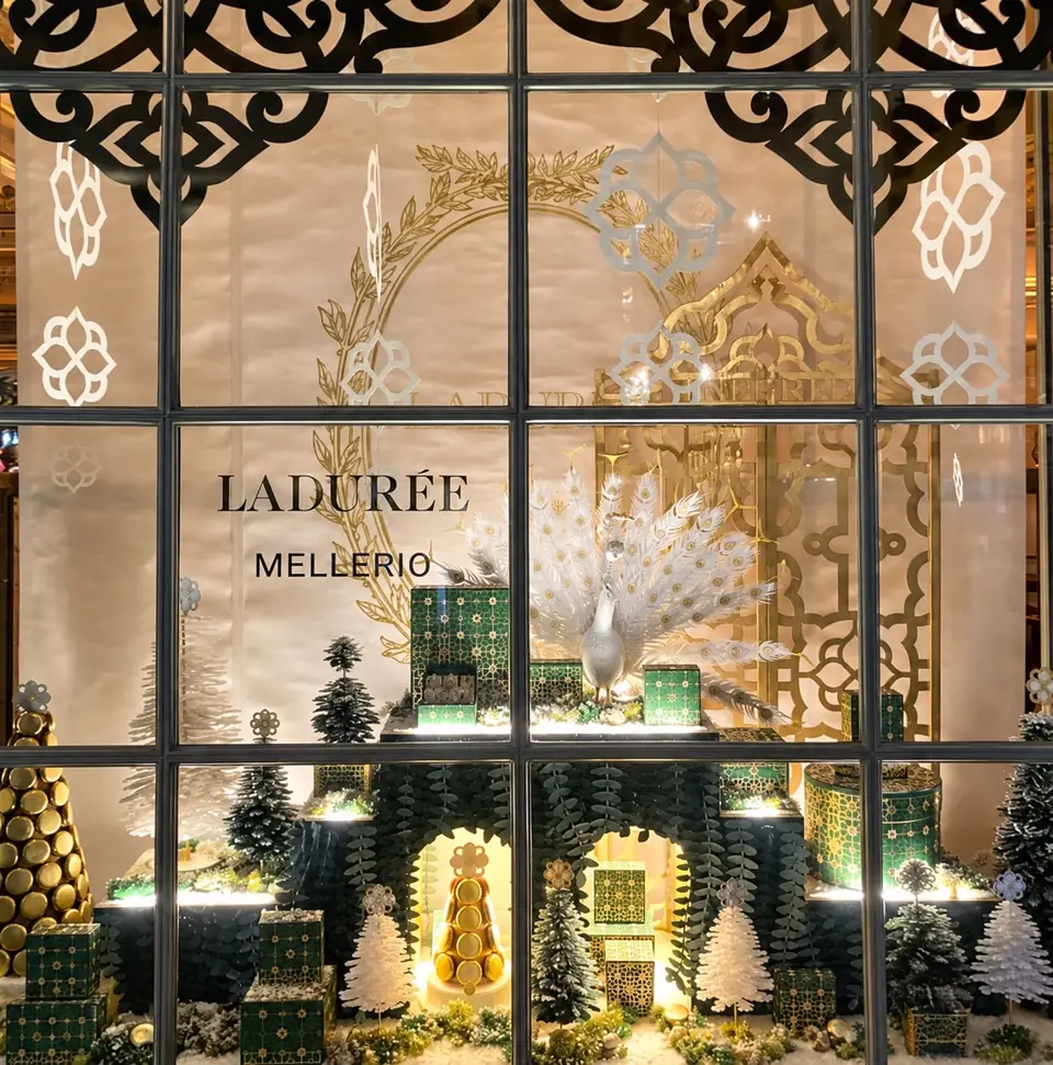 Vitrine de Noël Ladurée x Mellerio : arabesques dorées en vitrophanie, paon blanc et sapins en volume.