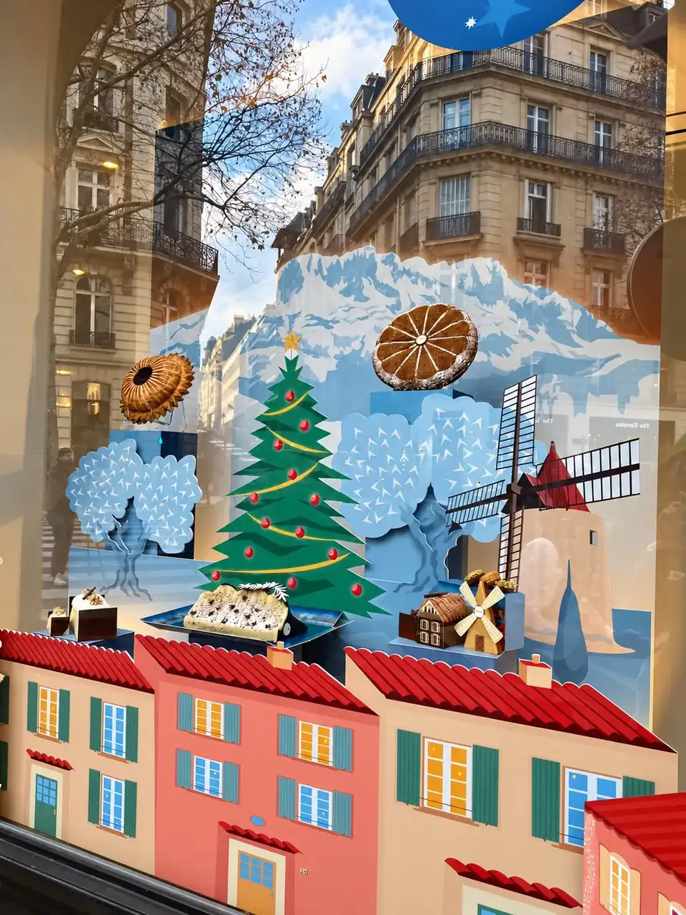 Vitrine Lenôtre « Un Noël en Provence » : village provençal, montagnes enneigées et sapin en impression découpée, reflets de la rue parisienne.